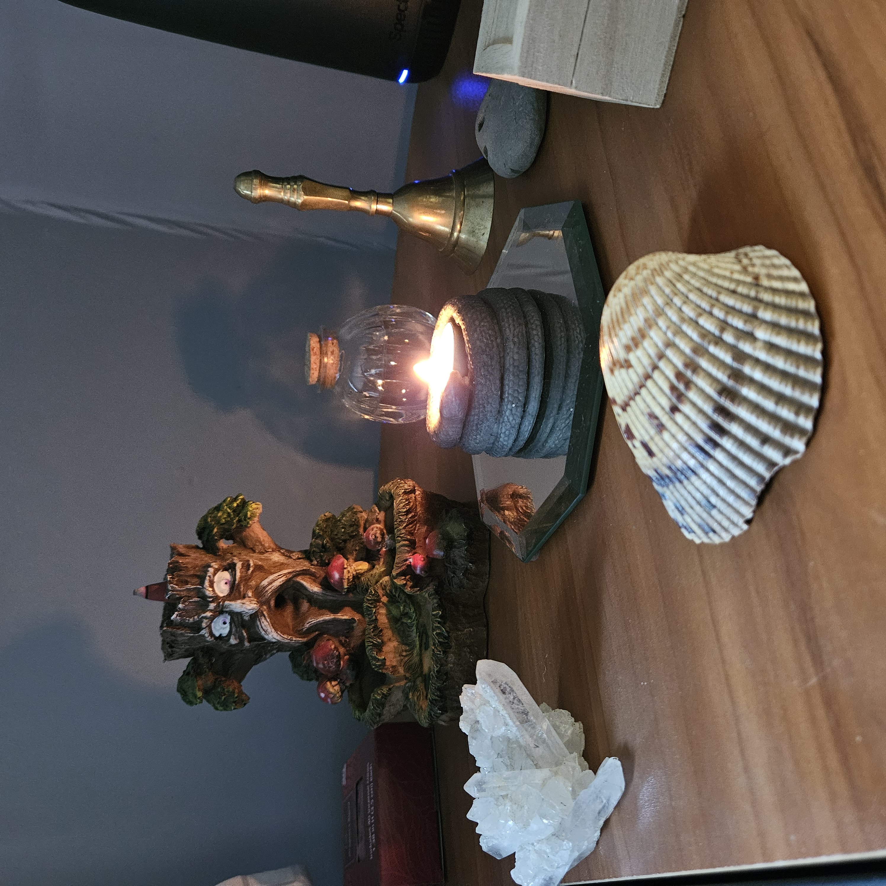 A lit candle sits in a candle holder that looks like a snake. Around the candle are various witchcraft objects including a seashell, a large quartz crystal, an incense burner, and a bell.